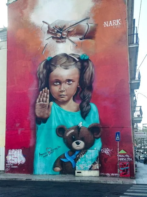 Mural of a girl holding a teddy bear and putting her hand up in a “stop” gesture. A hand is controlling puppet strings attached to her